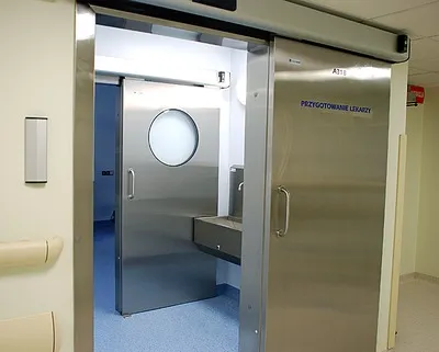 Puerta de acero robusta hospitalaria abierta, con vista al área de limpieza que incluye lavadero y acceso al quirófano, en cumplimiento con la NOM infraestructura y equipamiento de hospitales.